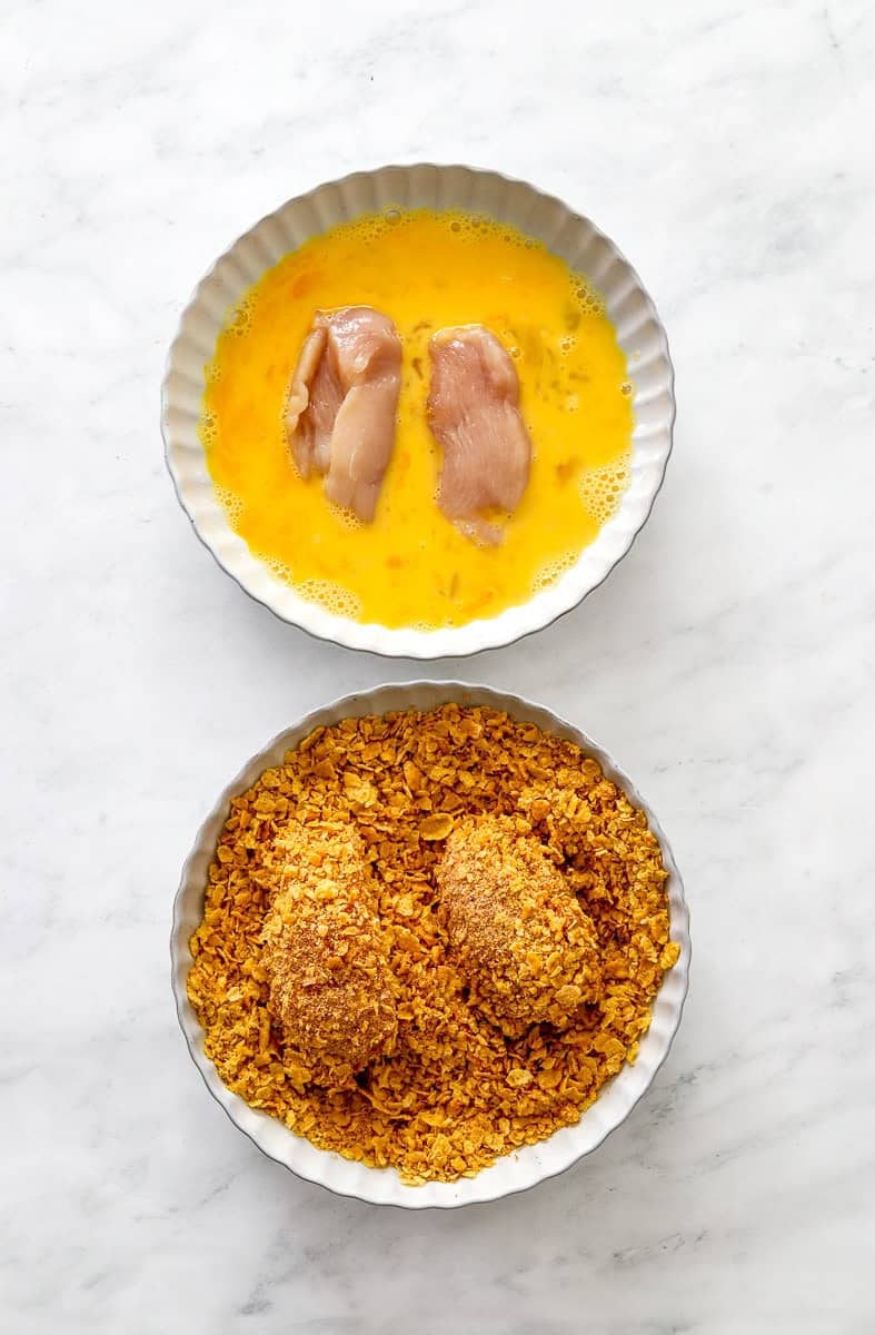 Bowl of whisked egg with two pieces of cut raw chicken in it with another shallow bowl fill with breading with coated chicken in the bowl with it. 