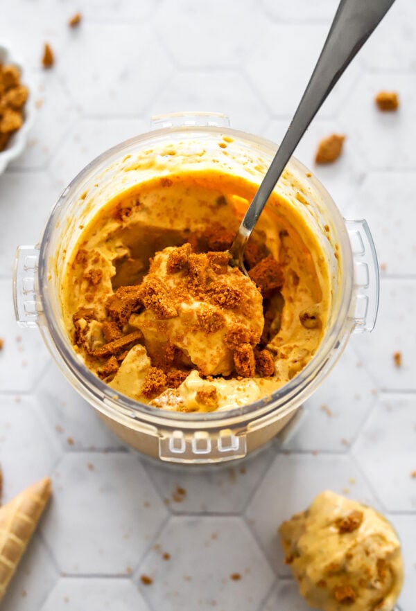 Container filled with healthy pumpkin protein ice cream with crushed Biscoff cookies with a spoon in the container with it and more ingredients around it.