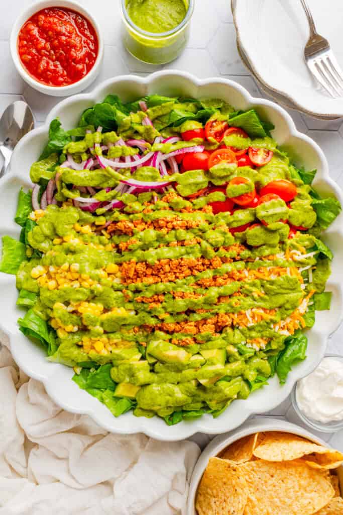 Taco salad with ground chicken in a salad bowl topped with green cilantro dressing with more ingredients around it. 