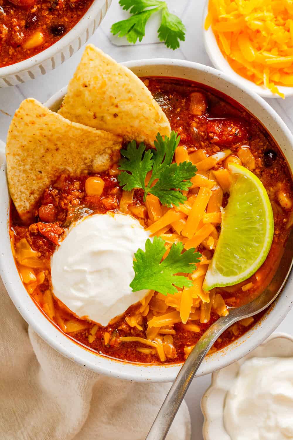 White bowl of instant pot healthy turkey chili topped with a couple tortilla chips, sour cream and cheddar cheese. 