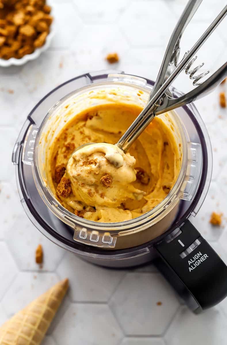 Protein pumpkin ice cream in Ninja Creami pint container with crushed Biscoff cookies in it with an ice cream scoop in the dish with it.