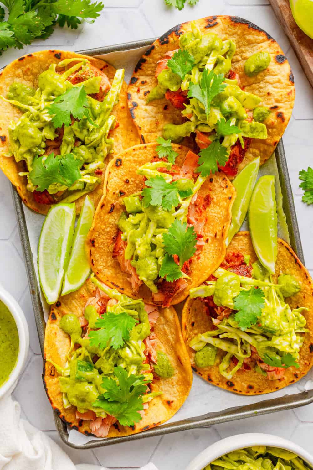 Tray filled with stuffed salmon lime tacos on corn tortillas with limes on the tray and cilantro leaves on the tacos.