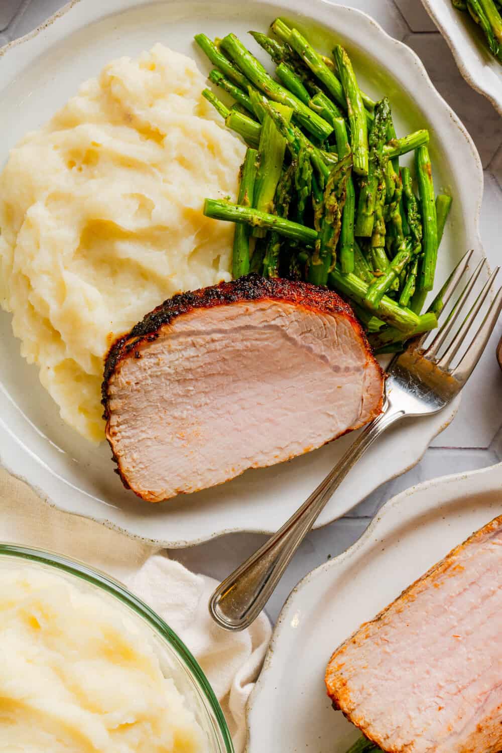Plate with a slice of pork loin roast on it with mashed potatoes and cut cooked asparagus on the the plate with it with more mashed potatoes in a bowl in front of it. 