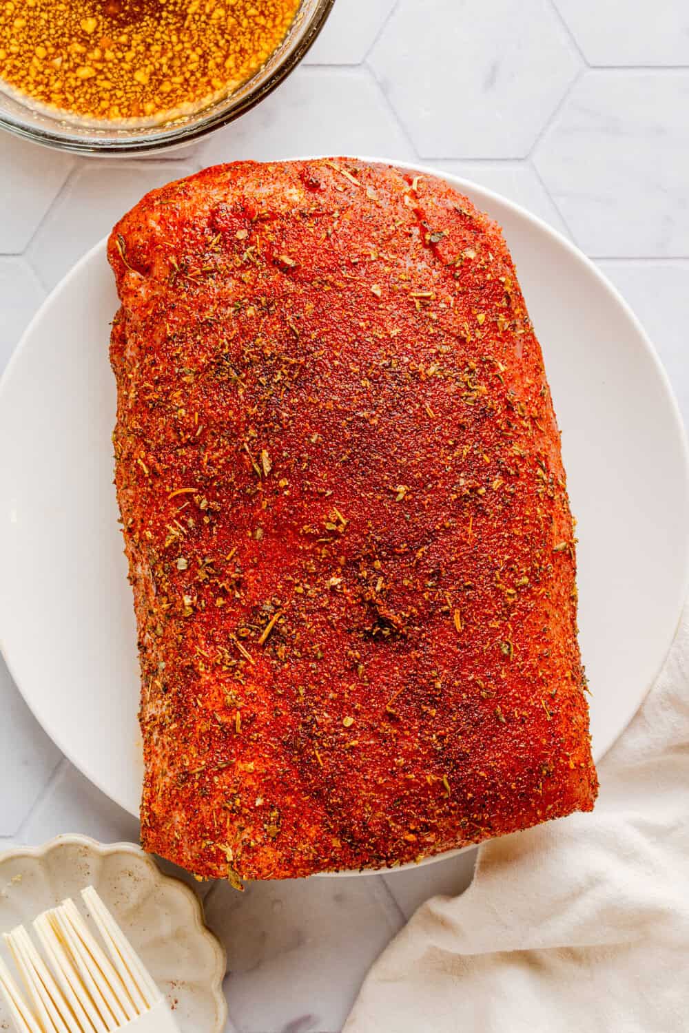 Uncooked pork loin roast on a plate with dry rub on it with more dry rub in a bowl behind it. 