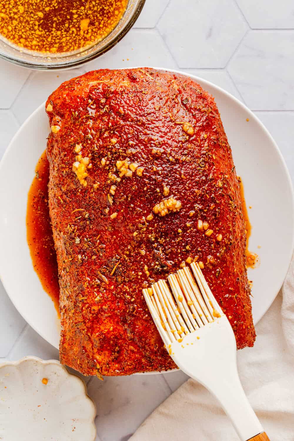 Raw pork loin on a plate with seasoning and glaze being brushed all over it. 