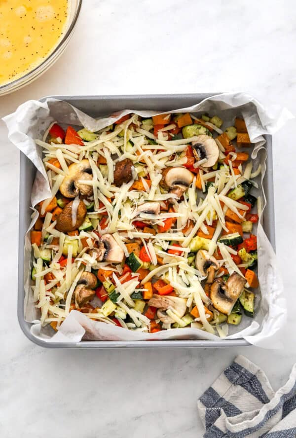 Roasted veggies and shredded cheese in the bottom of a lined pan with whisked eggs in a bowl behind it.