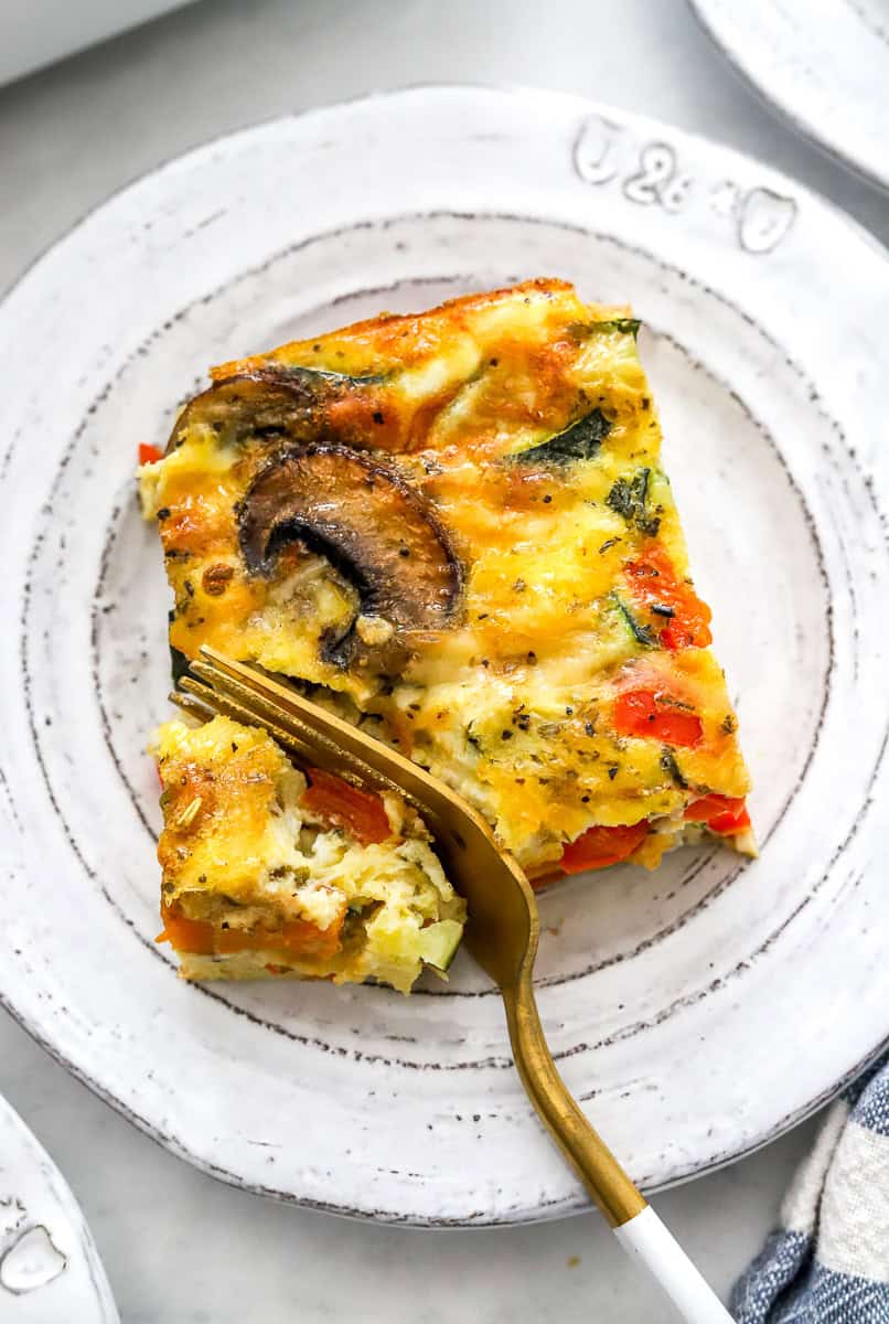 Square slice of baked vegetable frittata on a plate with a fork cutting into it.