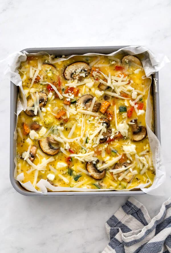 Unbaked frittata mixture in a square pan with a striped towel in front of it.