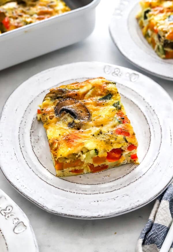 Square slice of veggie frittata on a round plate with more behind it.